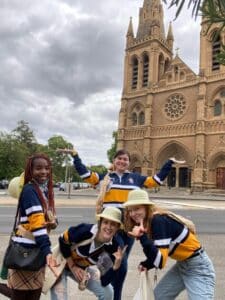 Four young women in Lincoln College rugby tops pose for a silly photo in front of St Paul's cathedral in Adelaide.